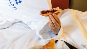 A person in a white robe holding a slice of toast with jam, sitting at a table with a basket of pastries, a plate with a fruit salad, and a glass of juice.