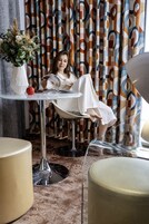 A person seated at a round glass table with a white cloth, reading a book. The room has a modern design with a patterned curtain in the background and a vase with flowers on the left.