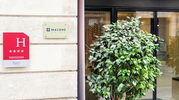 L’entrée d’un hôtel, avec une enseigne indiquant « MALONE HOTELS » et un panneau rouge en forme de « H » signalant la présence d’un hôtel.