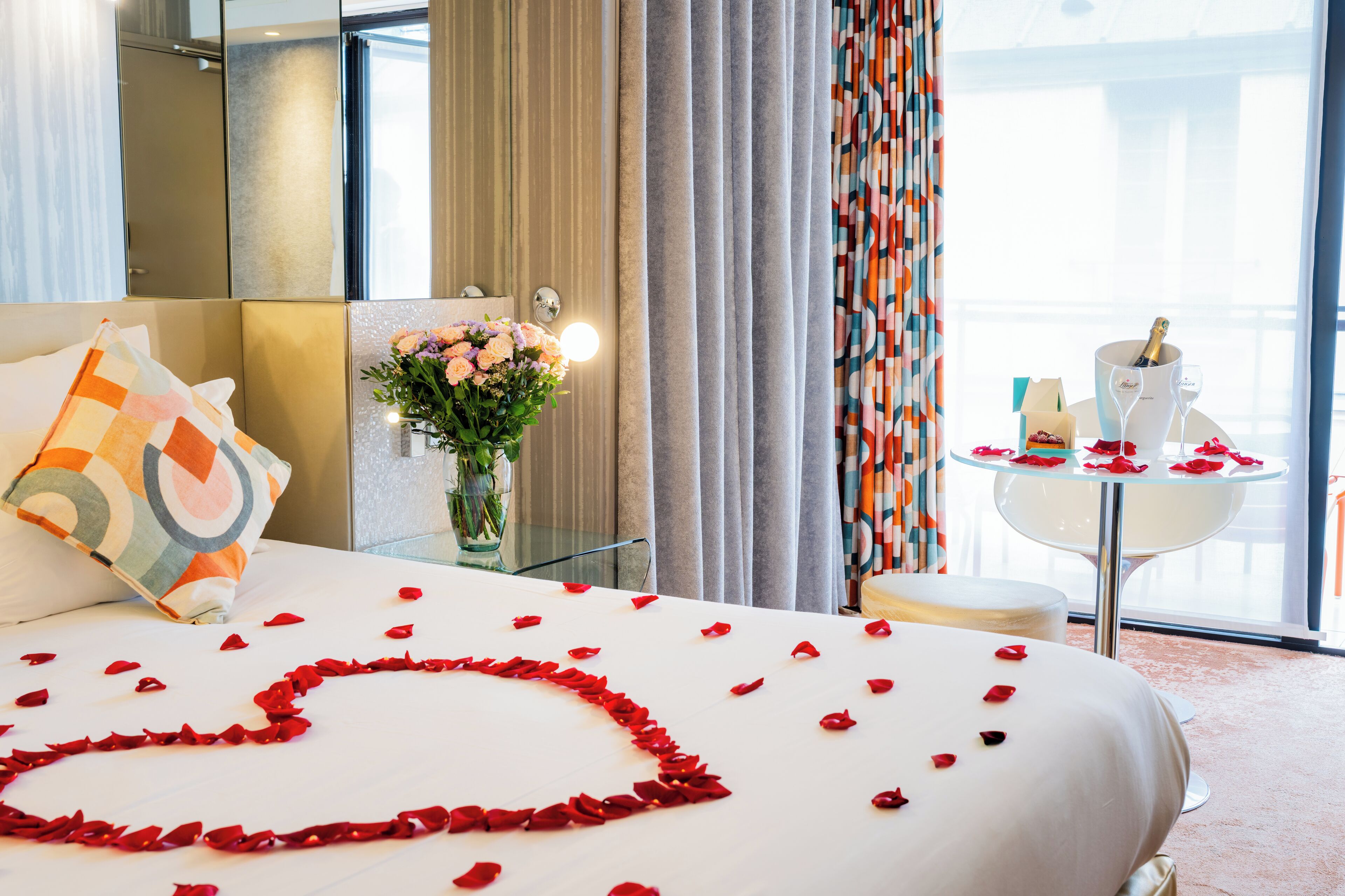 Een hotelkamer met een bed, een bloevas, een tafel met een fles en glas, en gordijnen.