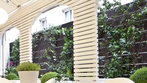 Een modern buitenzitgedeelte met witte tafels en stoelen, omringd door groen en een houten pergola.