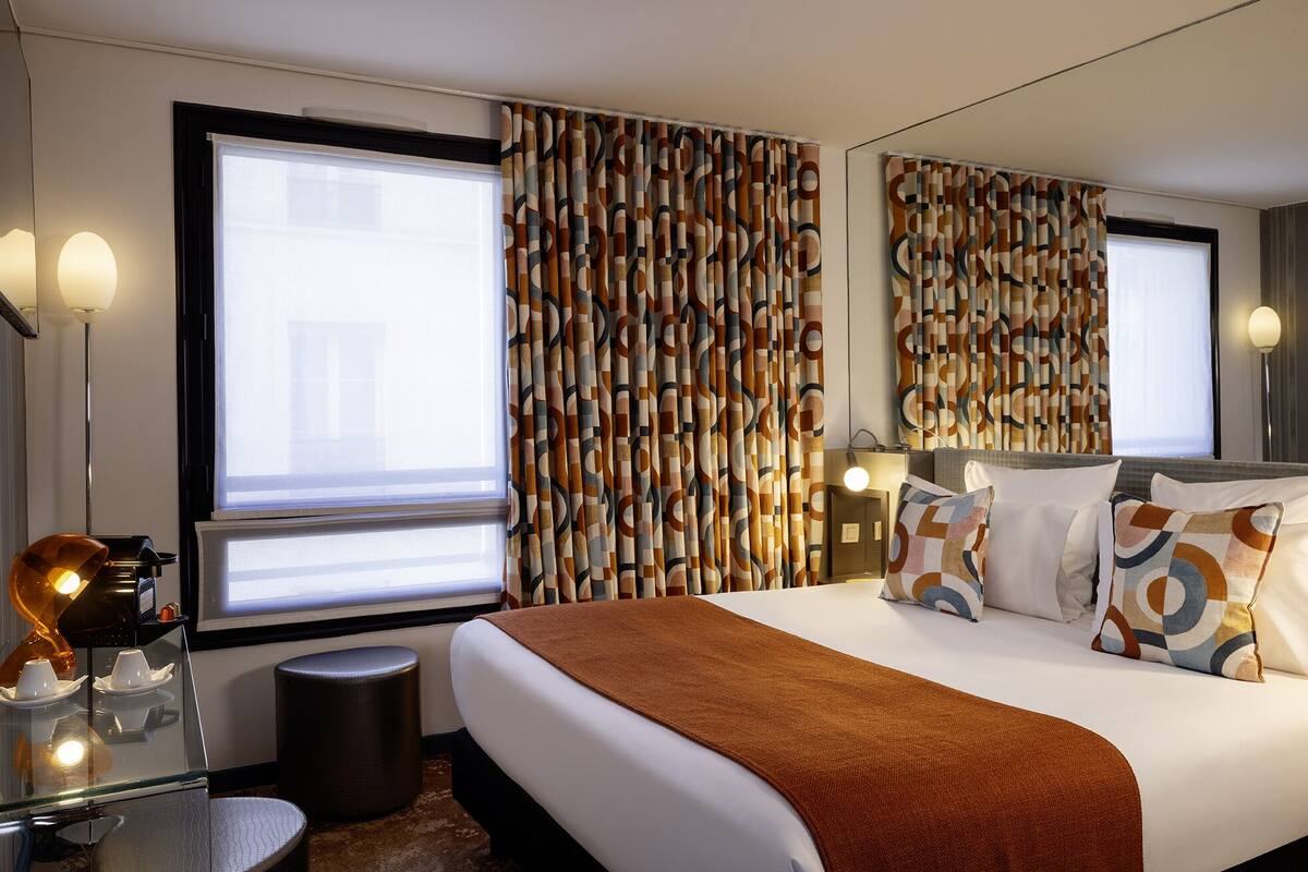 A hotel room with a large bed, a bedside table with a lamp, and patterned curtains.