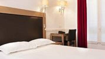 A hotel room with a large bed, a desk, two wall-mounted lamps, and a window with red curtains.