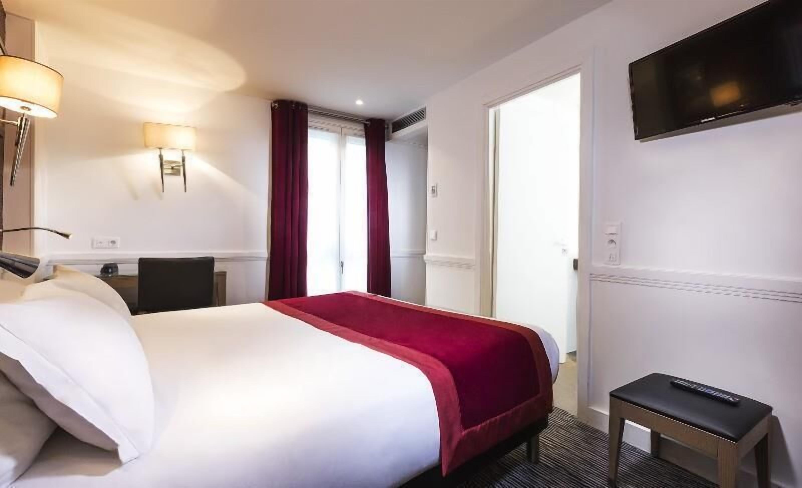 A hotel room with a bed, a desk, a chair, a TV mounted on the wall, and a door leading to another room.