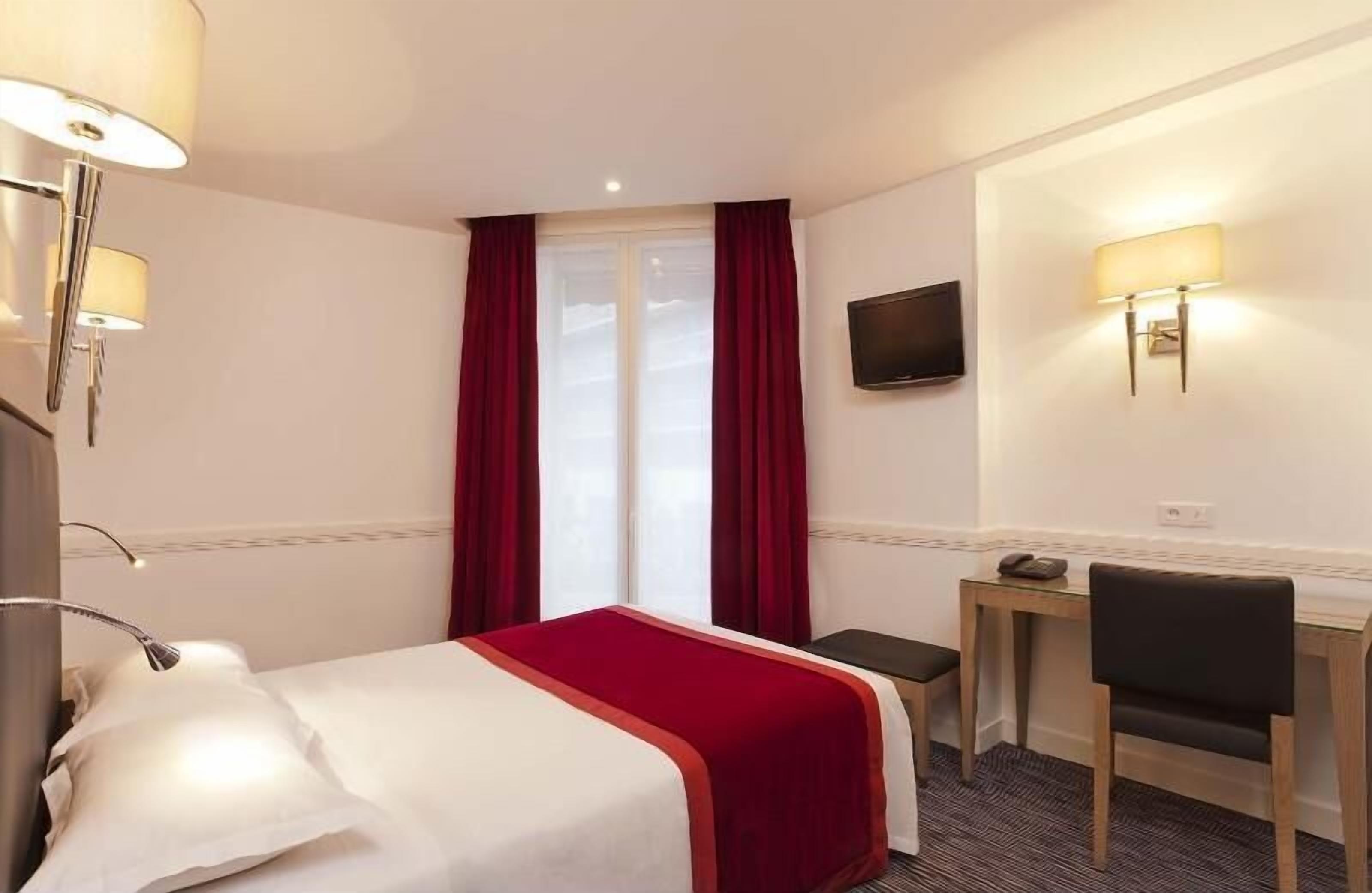 Habitación de hotel con cama, escritorio, silla, televisor y ventana con cortinas.