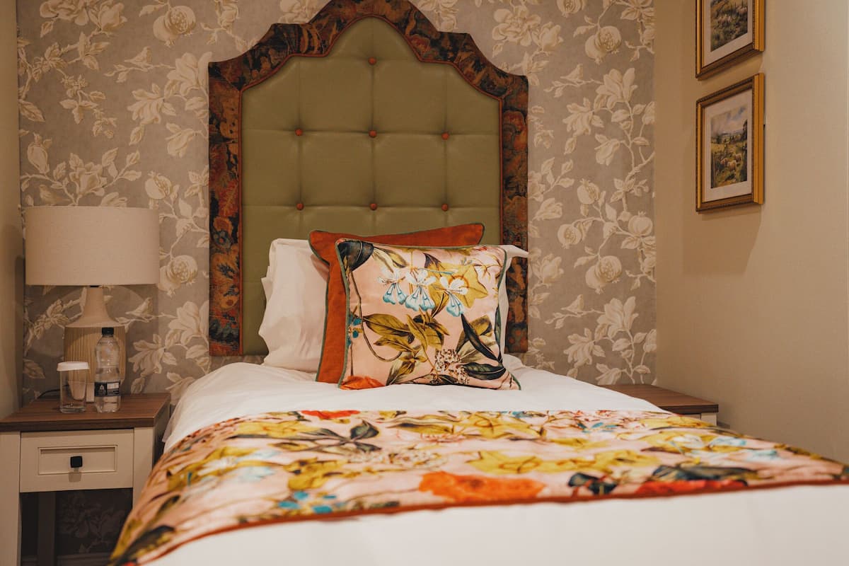 A neatly made bed with floral bedding, a green upholstered headboard, and a nightstand with a lamp and a bottle.