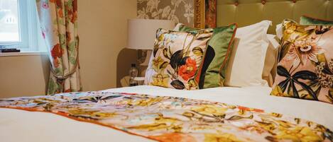 Una habitación de hotel con una cama, almohadas estampadas con motivos florales, un cabecero tapizado verde y una ventana con cortinas.