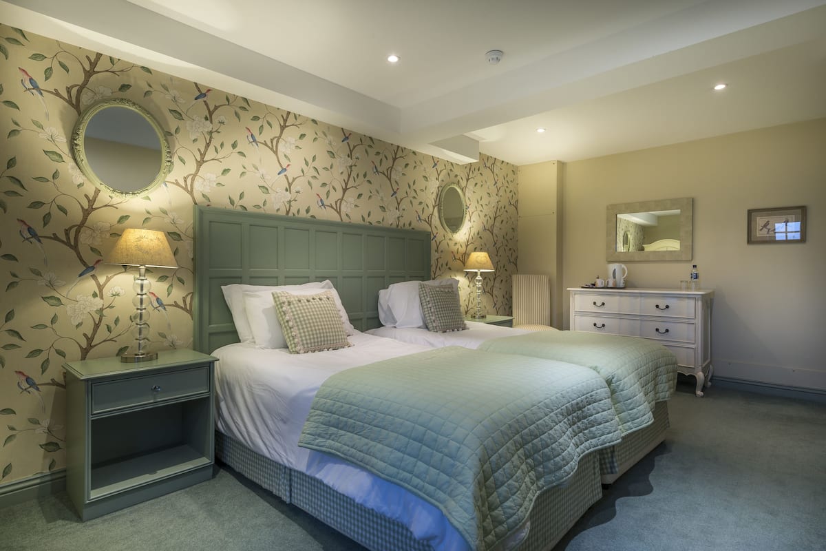 A bedroom with two beds, nightstands with lamps, a dresser, and floral wallpaper.