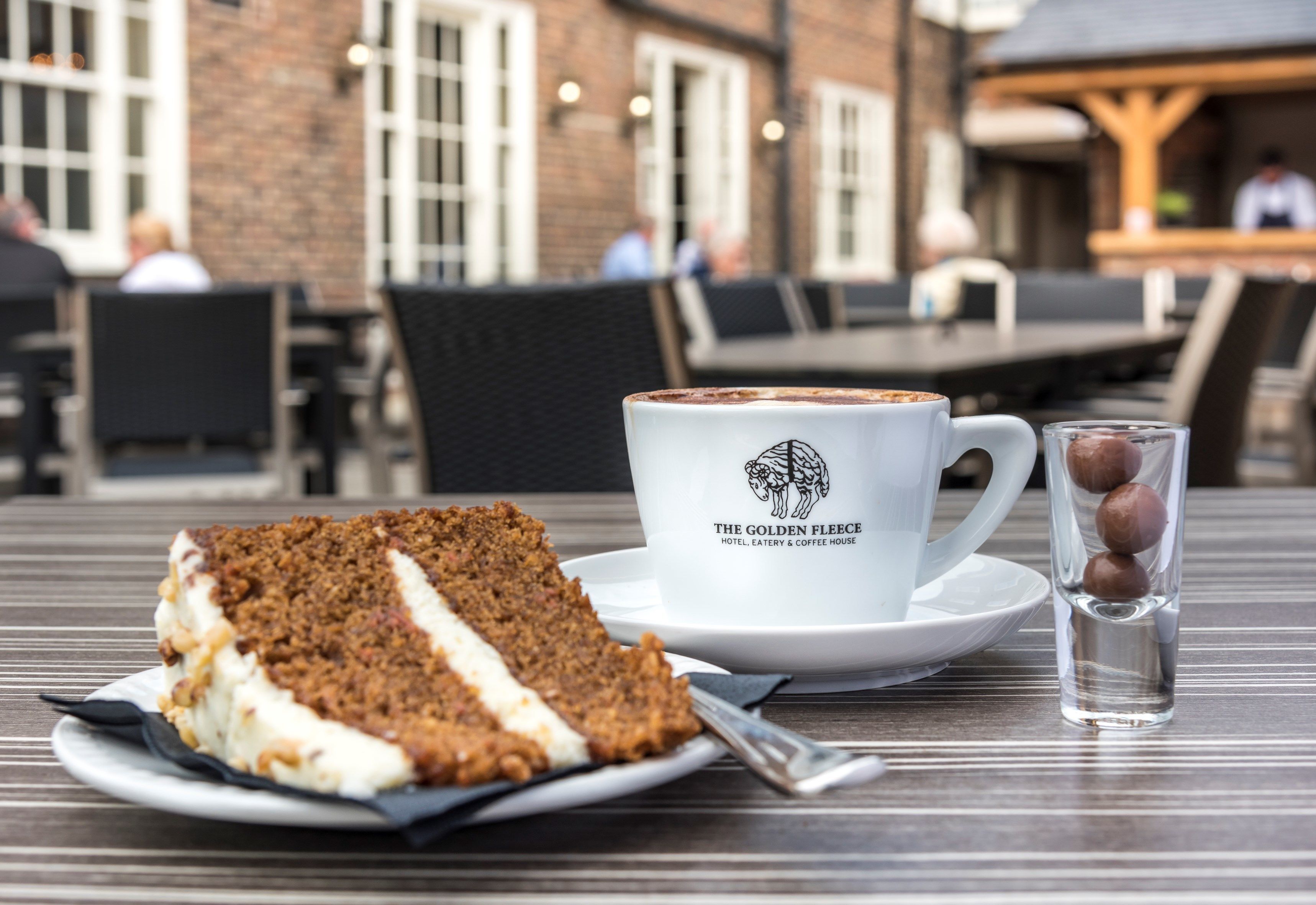 A slice of cake on a plate, a cup with a logo, and a glass with three round objects on a table in an outdoor setting with brick walls and people in the background.