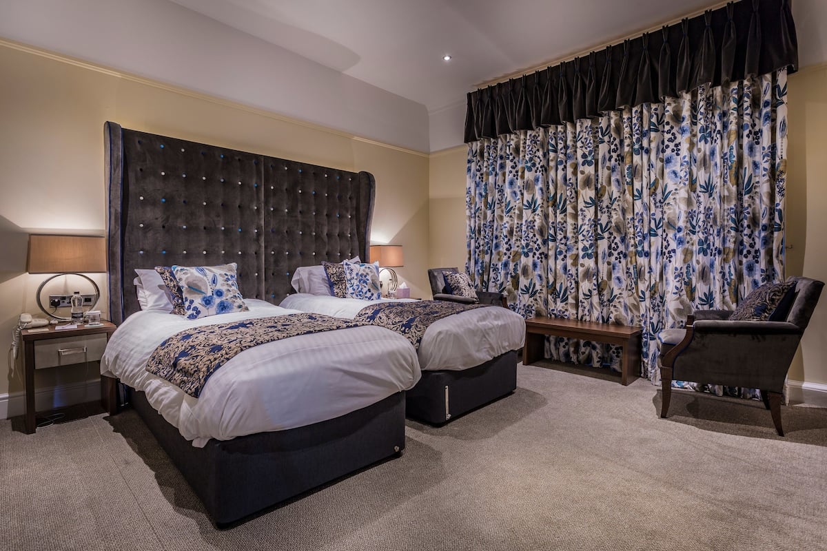 A hotel room with two beds, a large window with floral curtains, a bedside table with a lamp, and a chair.