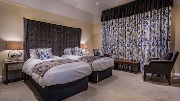 A hotel room with two beds, a large window with floral curtains, a bedside table with a lamp, and a chair.
