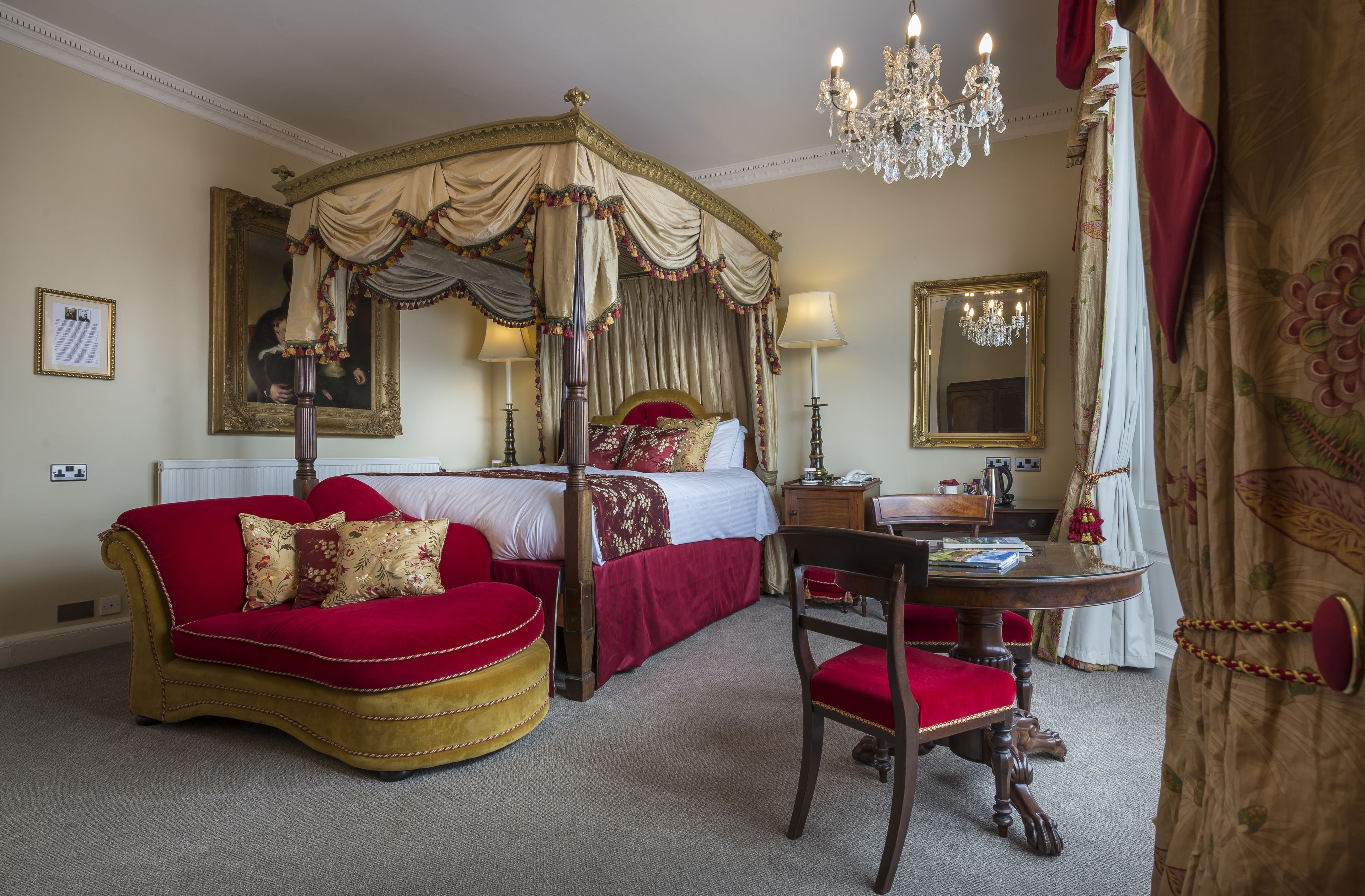 Una cama con dosel sobre cuatro columnas con una decoración en tonos rojo y dorado, un sofá tapizado en rojo, una silla de madera y una pequeña mesa redonda con una lámpara.