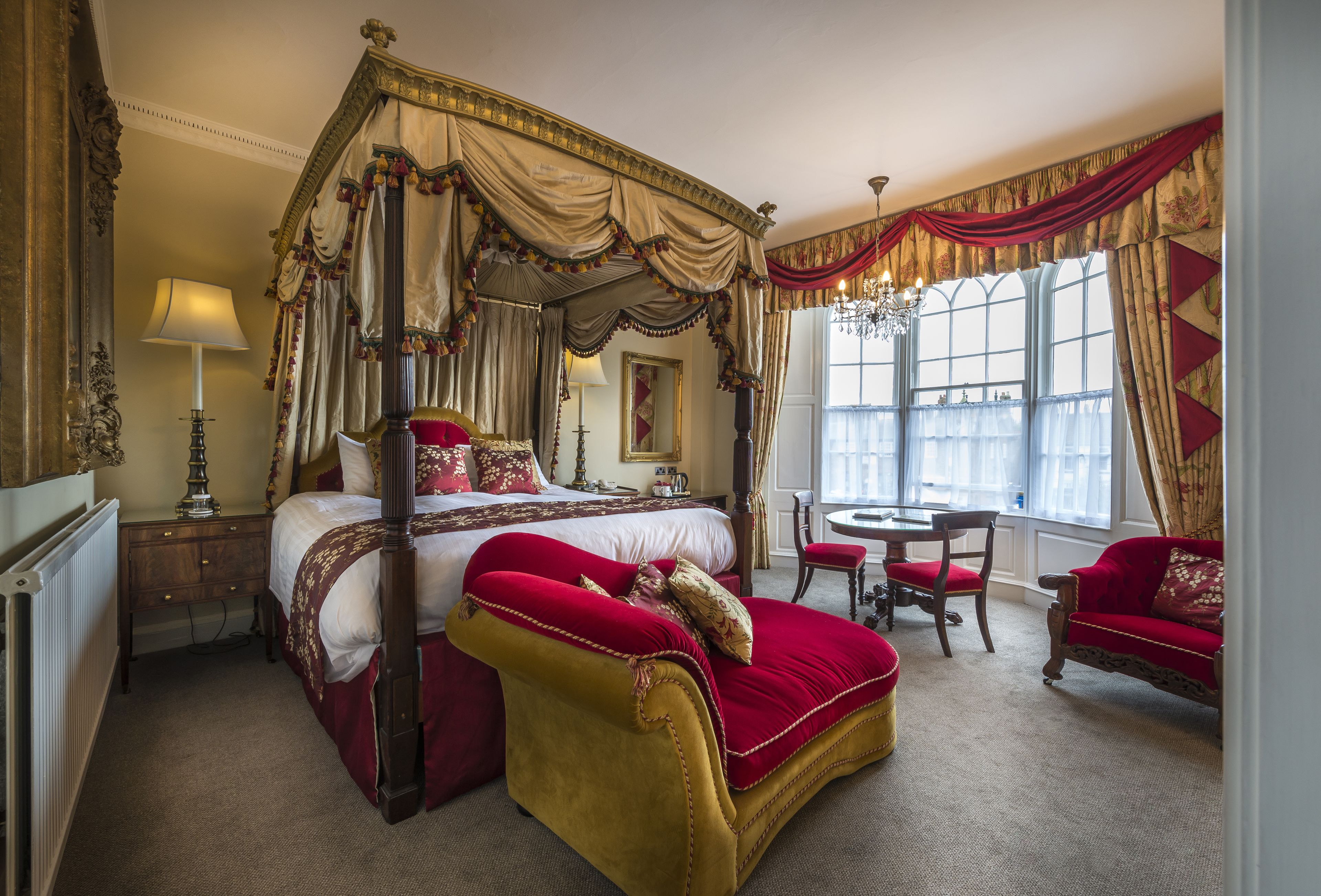 A luxurious bedroom with a four-poster bed, a sitting area with a red sofa, and a dining table.