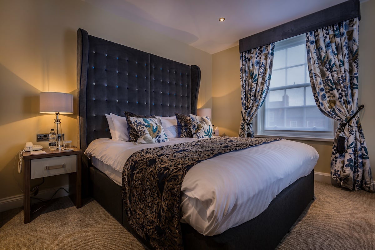 A hotel room with a large bed, a bedside table with a lamp, and a window with floral curtains.