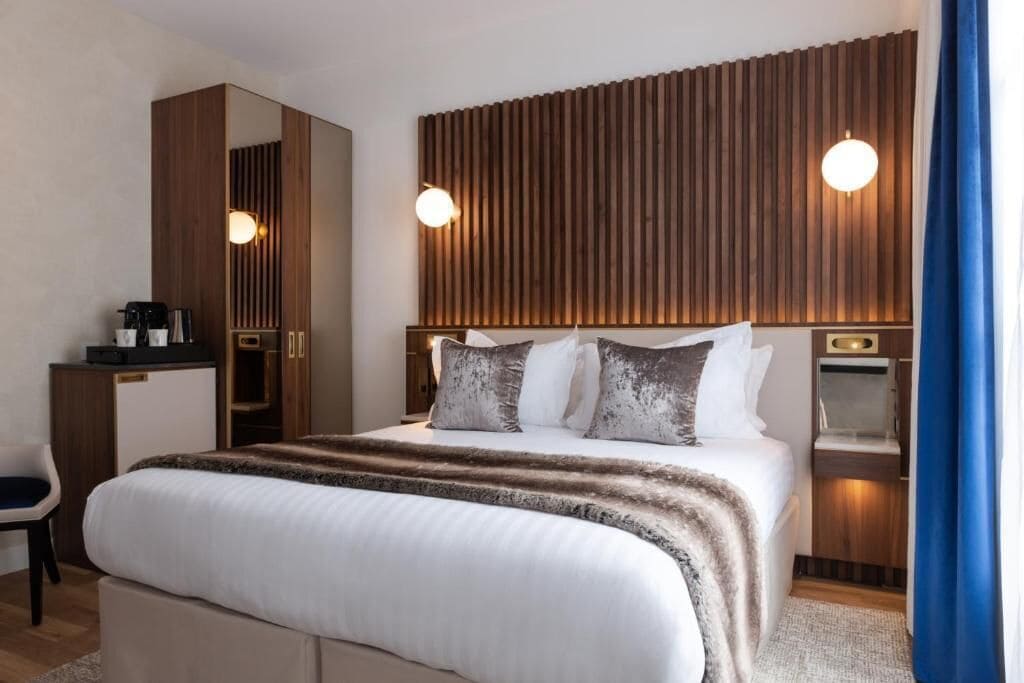 A modern hotel room with a large bed, a wooden headboard, bedside tables, a desk, and a mirror.