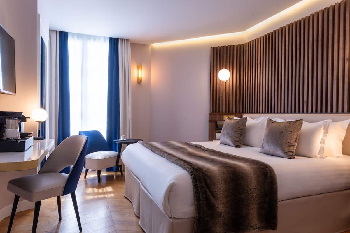 A modern hotel room with a large bed, a desk, a chair, and a view of the outdoors through large windows.