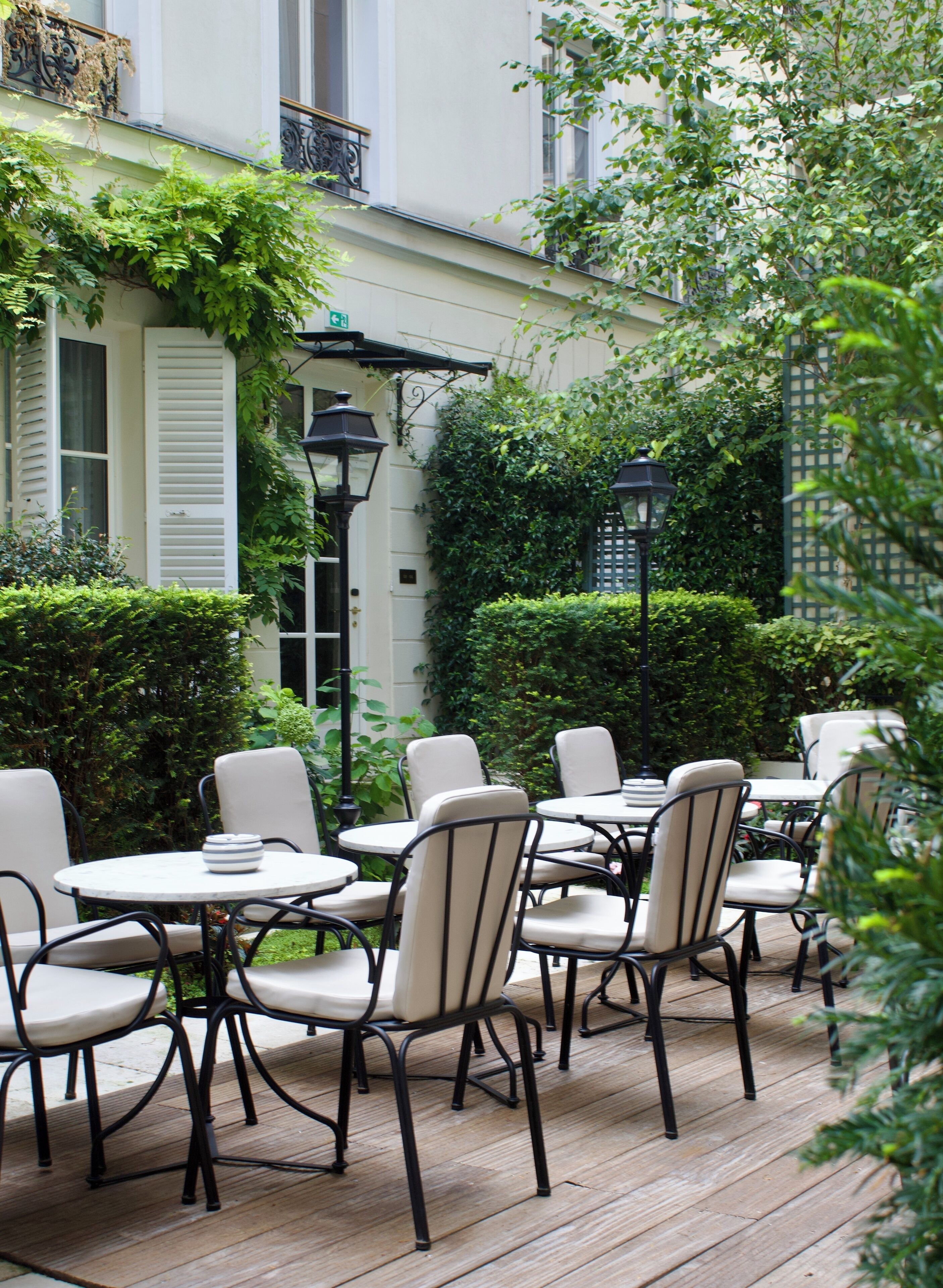An outdoor patio with round tables and chairs, surrounded by greenery and a white building with shutters.
