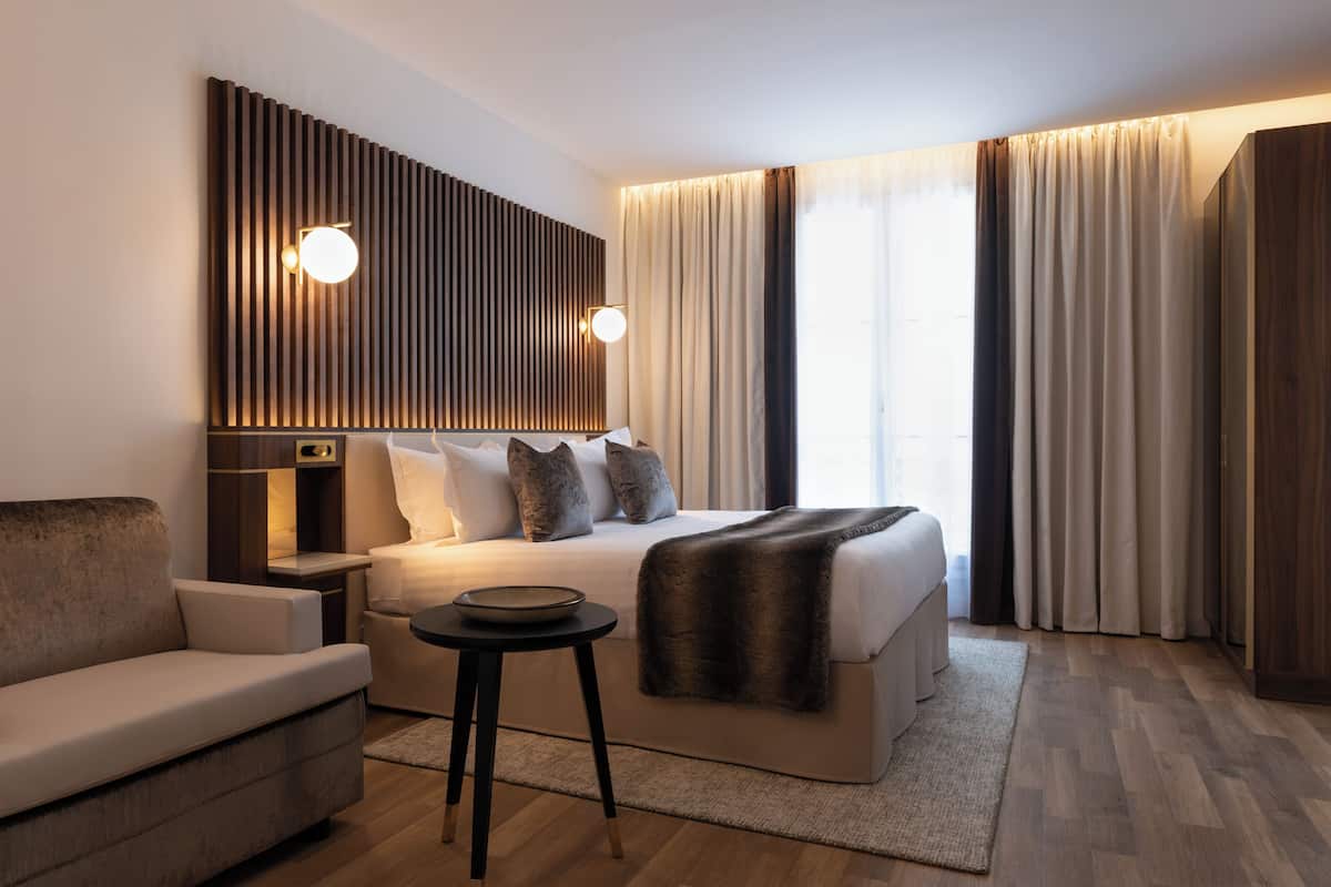A modern hotel room with a large bed, a sofa, a small table, and a wall-mounted light fixture.