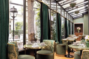 Un restaurante con sillas y mesas tapizadas de verde, ventanas amplias con cortinas y lámparas colgantes.