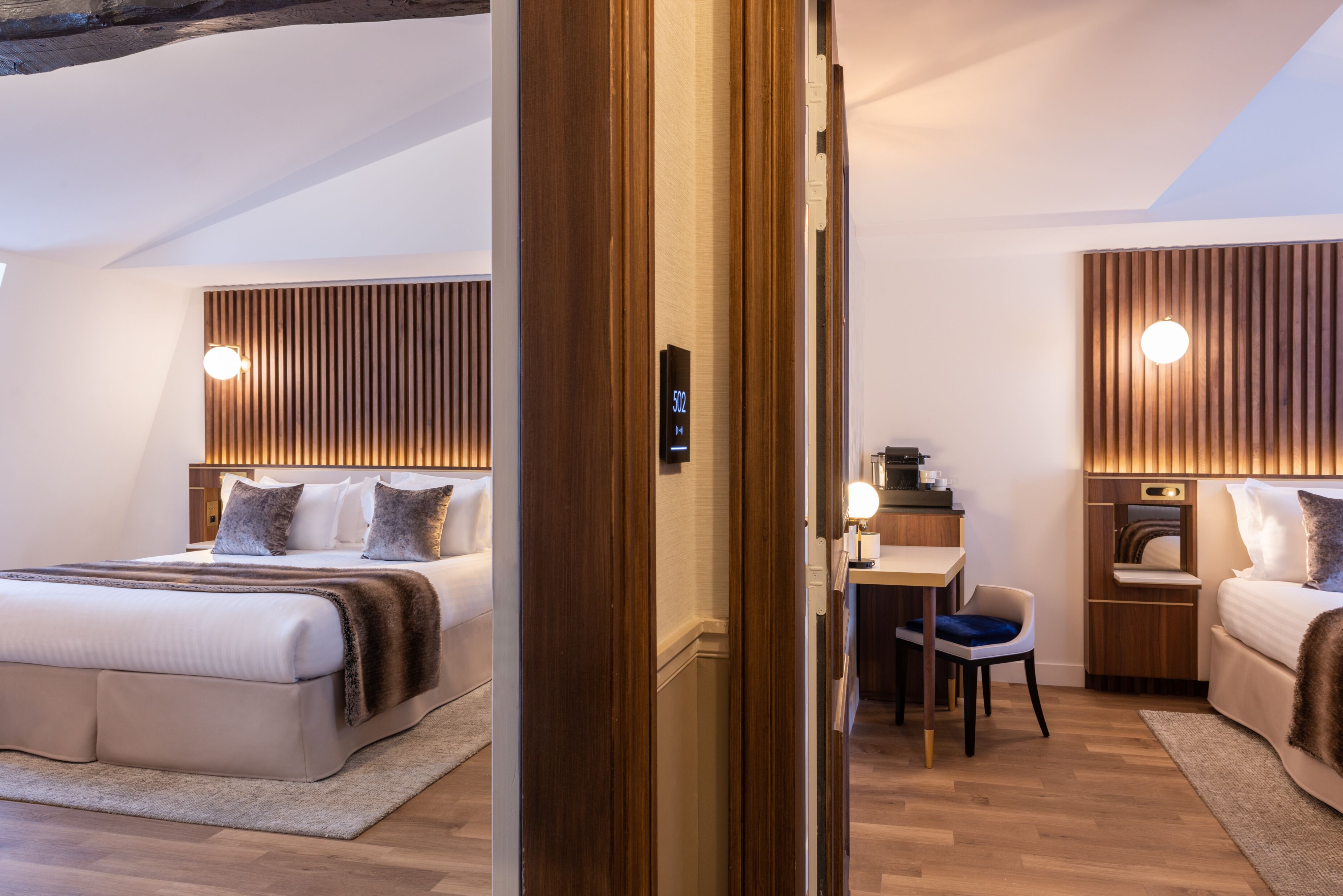 A modern hotel room with a large bed, a desk, and a chair. There is a wooden headboard and a wall-mounted light fixture.