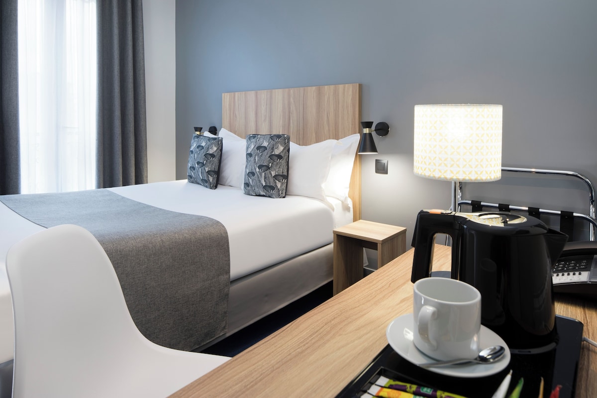 A hotel room with a large bed, a bedside table with a lamp, a coffee maker, and a cup on the table.