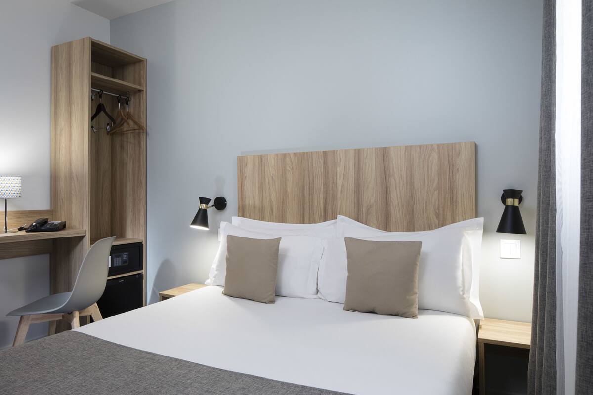 A modern hotel room with a large bed, a desk with a chair, a wardrobe, and bedside lamps.