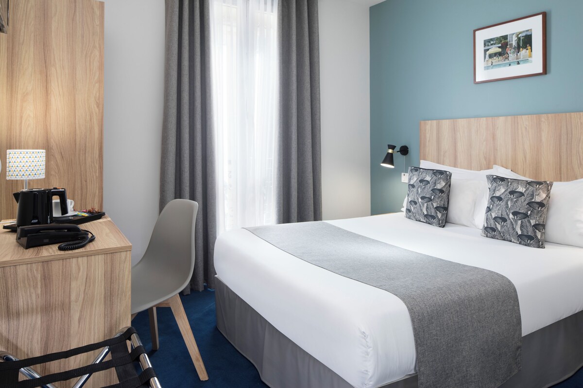 Een hotelkamer met een bed, een bureau met lamp, een stoel en een raam met gordijnen.