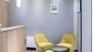 A reception area with a wooden counter, two yellow chairs, a small round table, and a framed picture on the wall. - Hotel Moulin Vert (Paris)