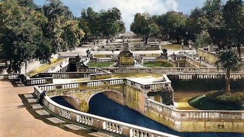 Un jardín amplio y ornamentado con una fuente central, rodeado de senderos de piedra y bordeado de árboles.