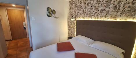 A hotel room with a bed, two pillows, a nightstand, a wall-mounted lamp, and a floral wallpaper.