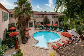 A pool area with lounge chairs and umbrellas, surrounded by a building with green shutters and a tiled roof.