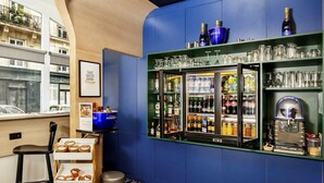 A bar area with a blue wall, shelves with bottles and glasses, a trash can, and a chair. - ibis Styles Paris Cadet Lafayette (Paris)