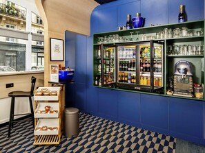 A bar area with a blue wall, shelves with bottles and glasses, a trash can, and a chair. - ibis Styles Paris Cadet Lafayette (Paris)