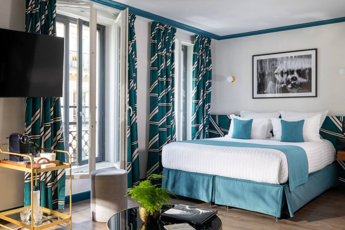 A hotel room with a bed, a TV, a window with curtains, a small table with glasses, and a plant.