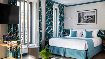 A hotel room with a bed, a TV, a window with curtains, a small table with glasses, and a plant.