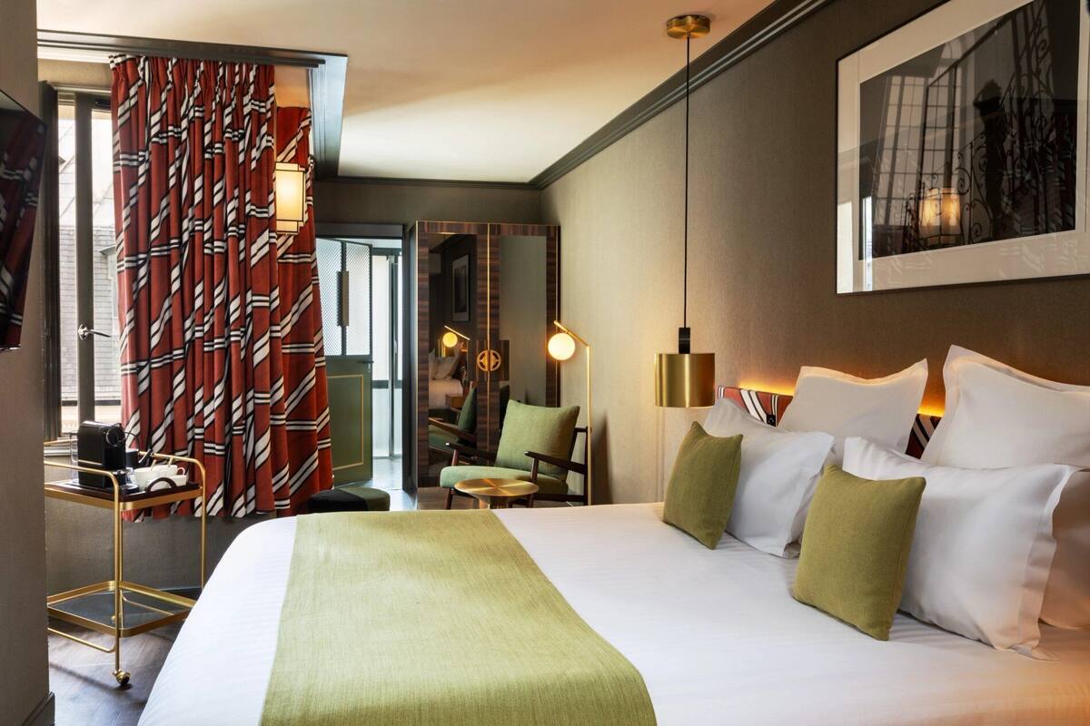 A hotel room with a large bed, a green throw, a side table, and a chair. There is a window with curtains and a view of the outside.