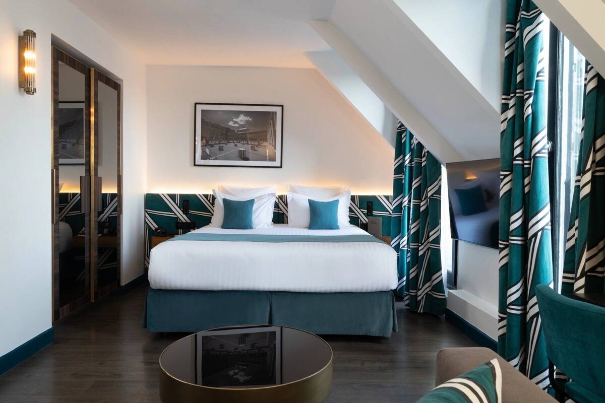 A modern hotel room with a large bed, a bedside table, a round coffee table, and a view of the cityscape through the window.