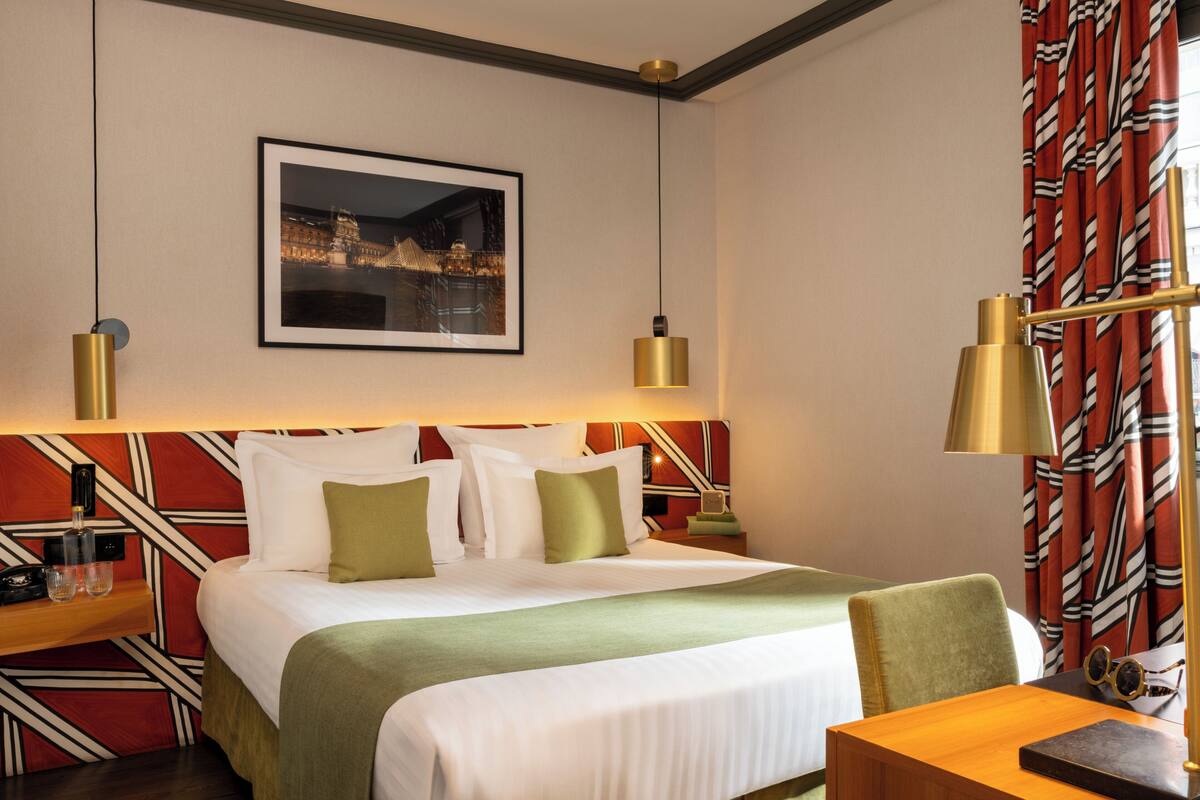 A hotel room with a bed, bedside tables, a desk, a chair, a framed picture on the wall, and a patterned curtain.