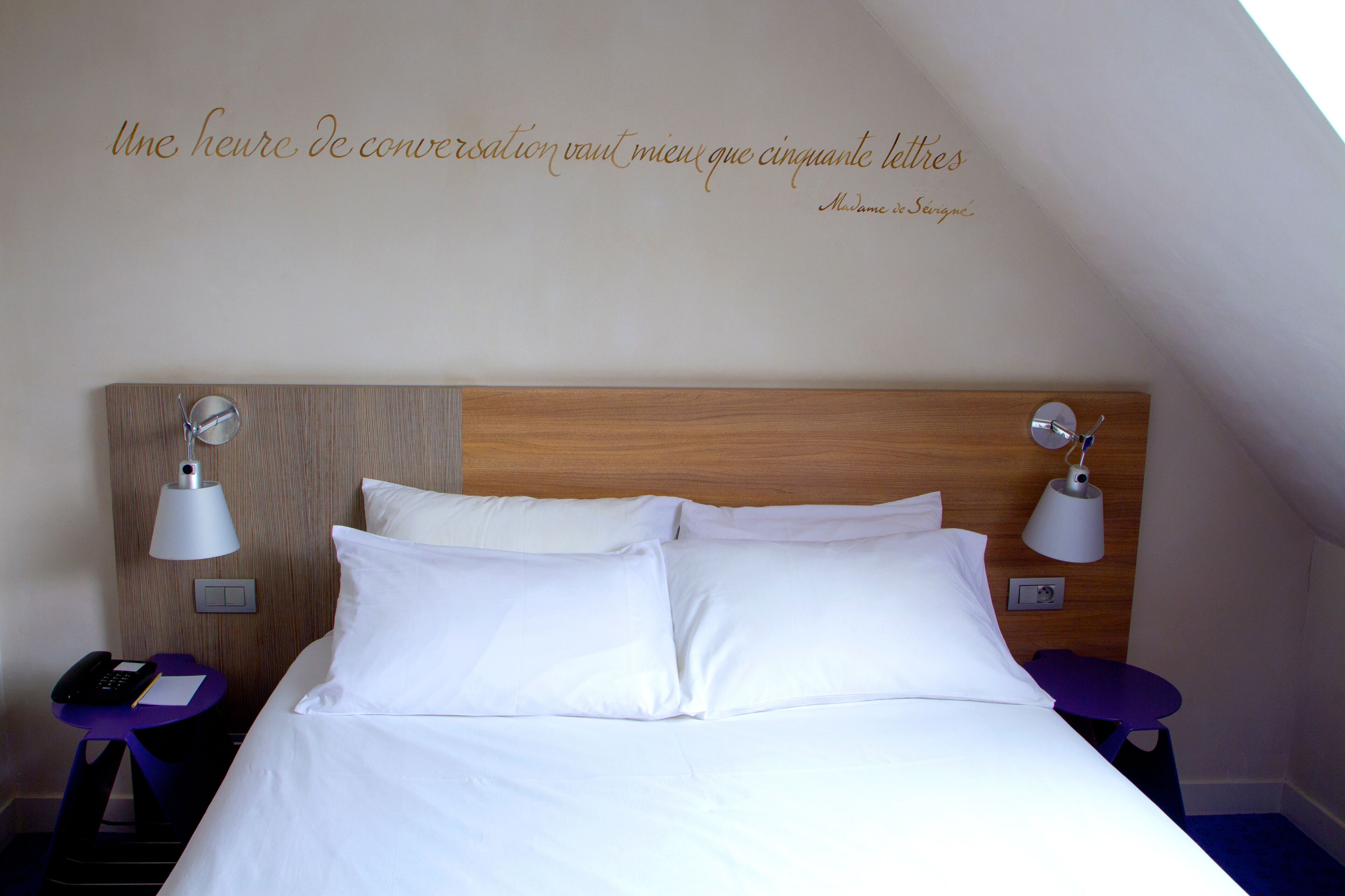 A hotel room with a wooden headboard, white bedding, two bedside lamps, a small table with a purple chair, and a wall-mounted light switch.