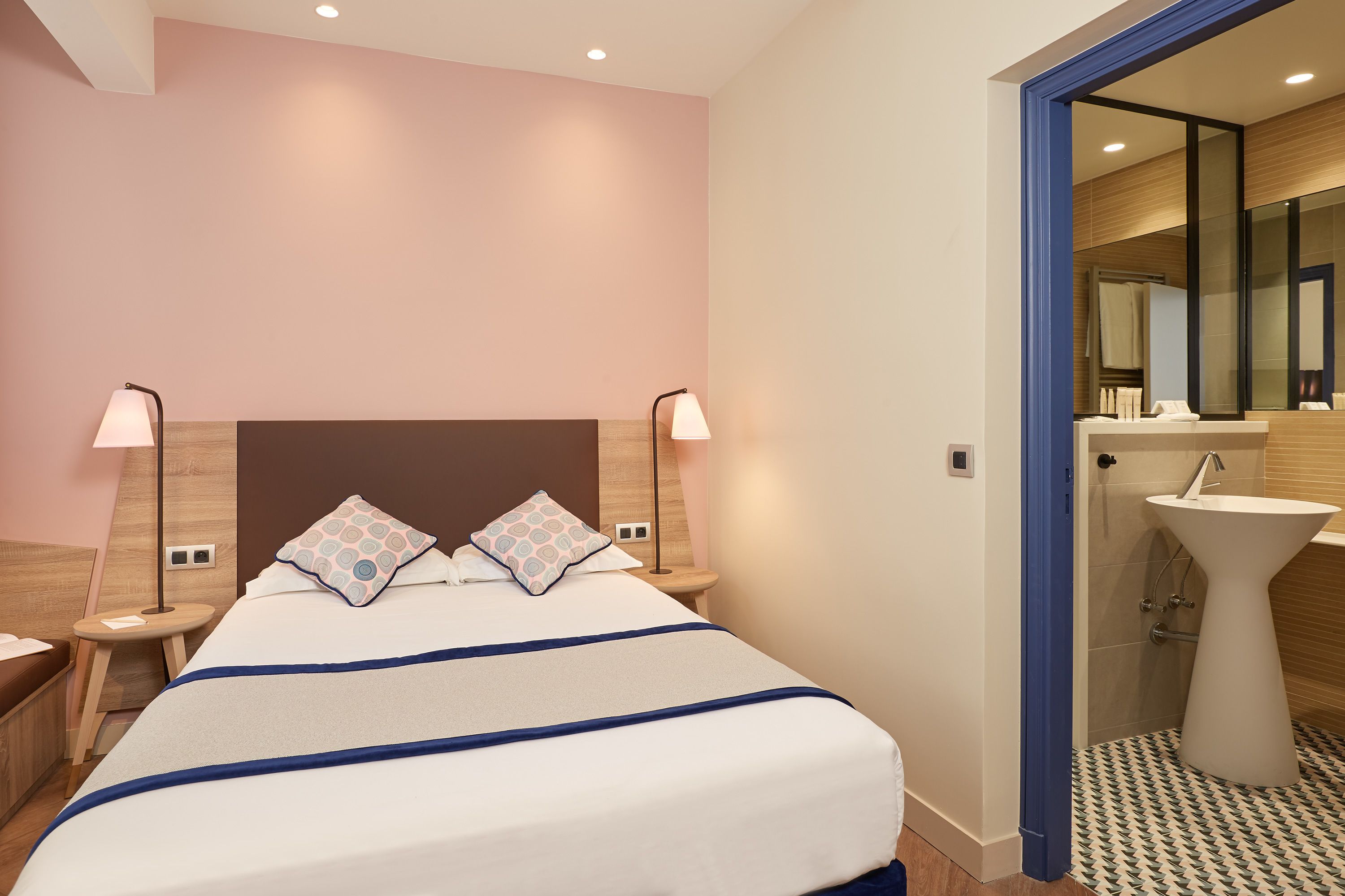 A hotel room with a bed, bedside lamps, a nightstand, a bathroom with a sink and mirror.