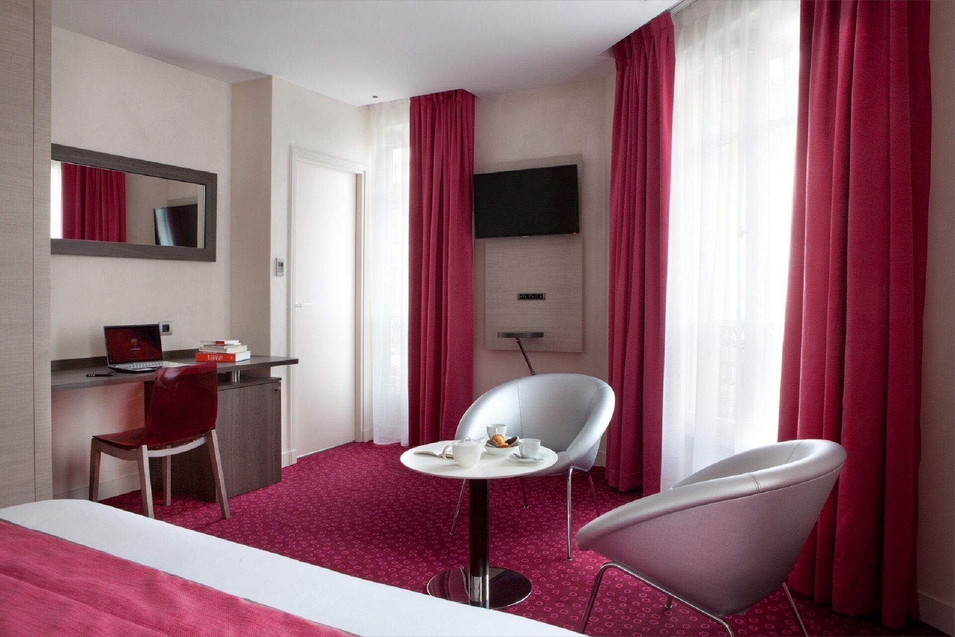 A hotel room with a bed, a desk with a laptop, a chair, a small table with a cup and a plate, and a TV mounted on the wall.