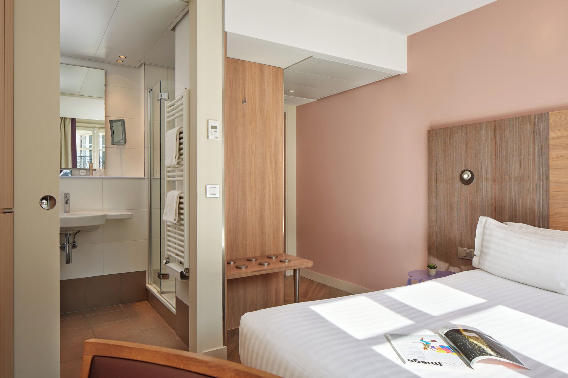 A hotel room with a bed, a bedside table with a magazine, a wooden wardrobe, a bathroom with a sink and mirror, and a towel rack.