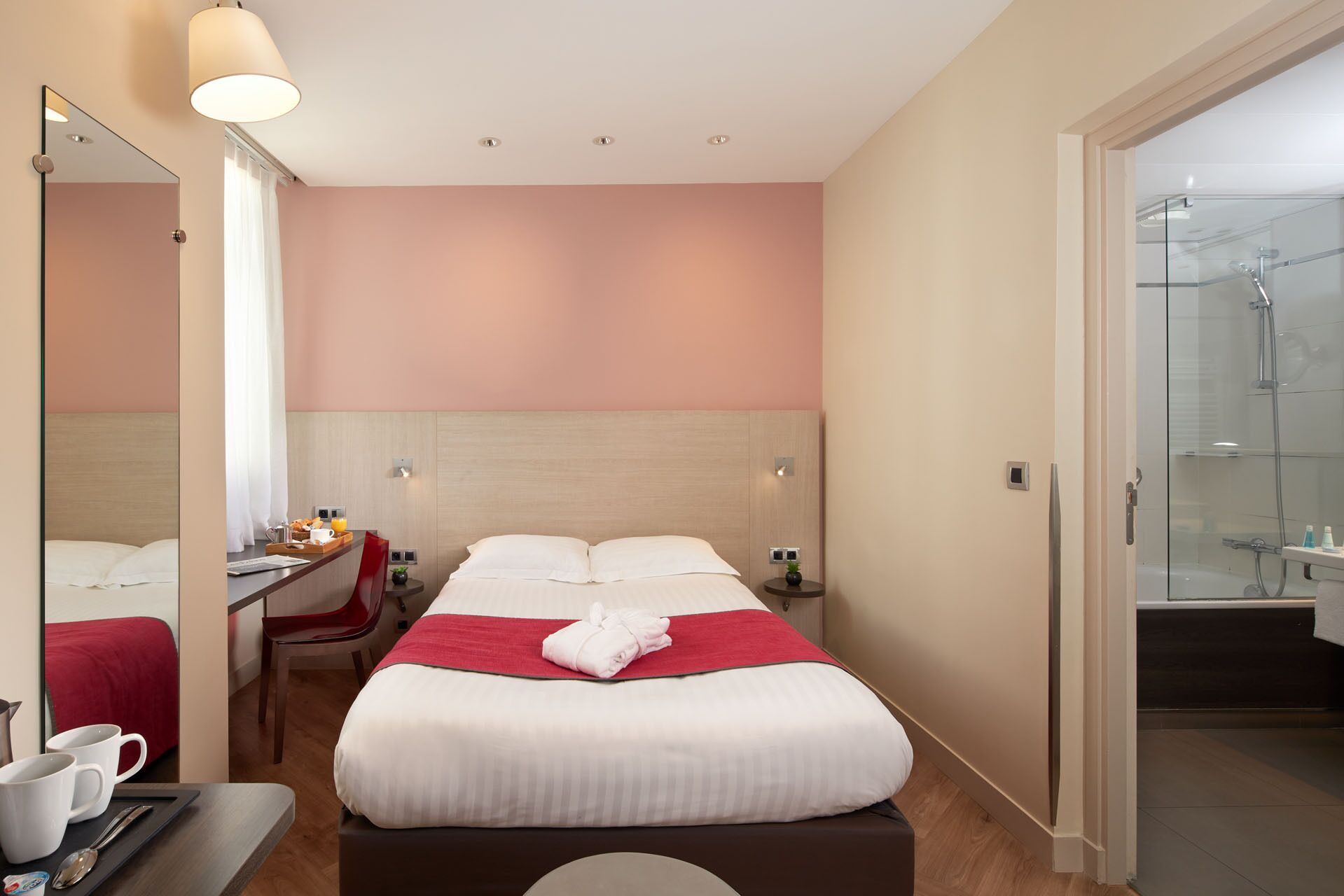 A hotel room with a bed, a desk, a chair, a small table with a red cloth, a mirror, and a shower area.