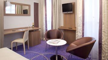 A hotel room with a desk, two chairs, a TV, a bed, and a window with curtains.