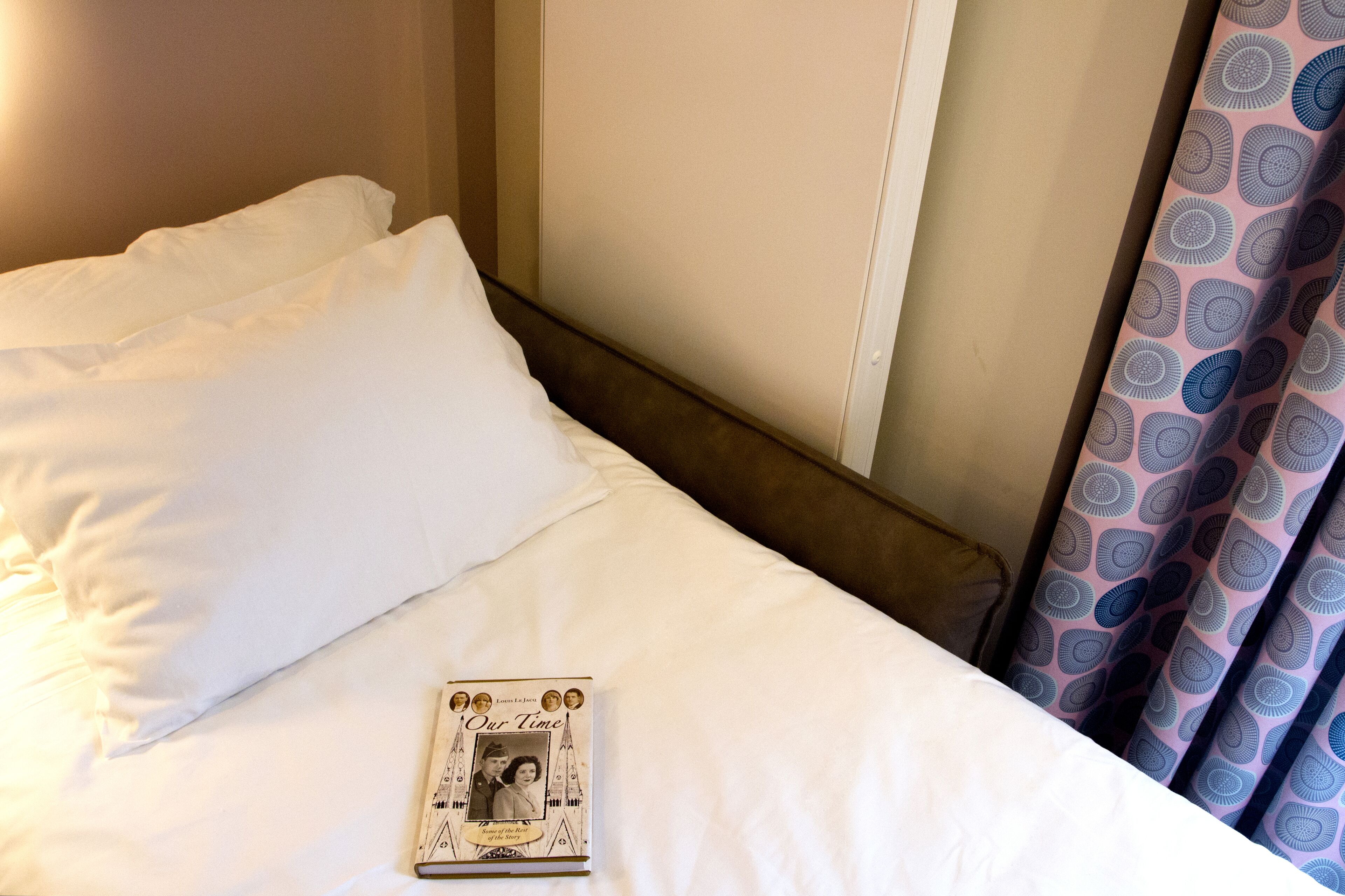 A neatly made bed with white linens, a book titled 'Our Time' on the bed, and a patterned curtain partially visible.