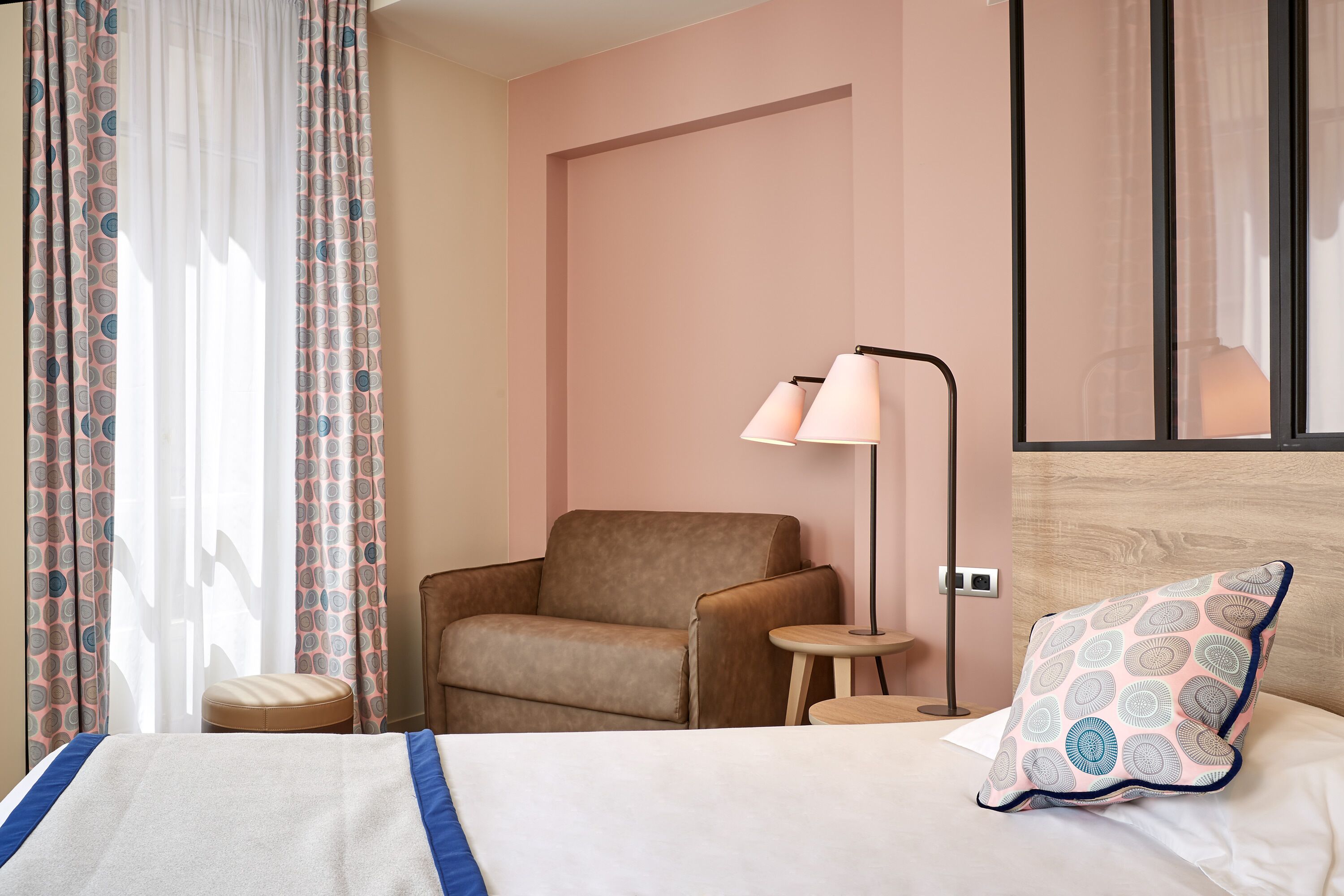 A hotel room with a bed, a chair, a small table, a lamp, and a patterned pillow.