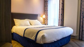 Una cama bien tendida con cabecera, lámpara de noche y ventana con cortinas estampadas.