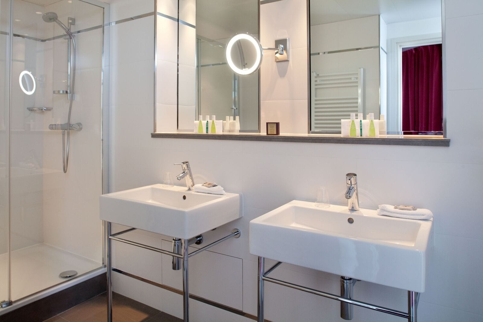 Une salle de bain moderne avec deux vasques, une douche à l’italienne en verre et un grand miroir avec éclairage intégré.