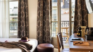A hotel room with a bed, a desk with a laptop, a coffee machine, and a view of the city through large windows. - Hotel de Sevigne (Paris)