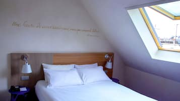 Una cama bien hecha con sábanas blancas, cabecera de madera y dos lámparas de noche. Junto a la cama, una mesita con un teléfono y una libreta.